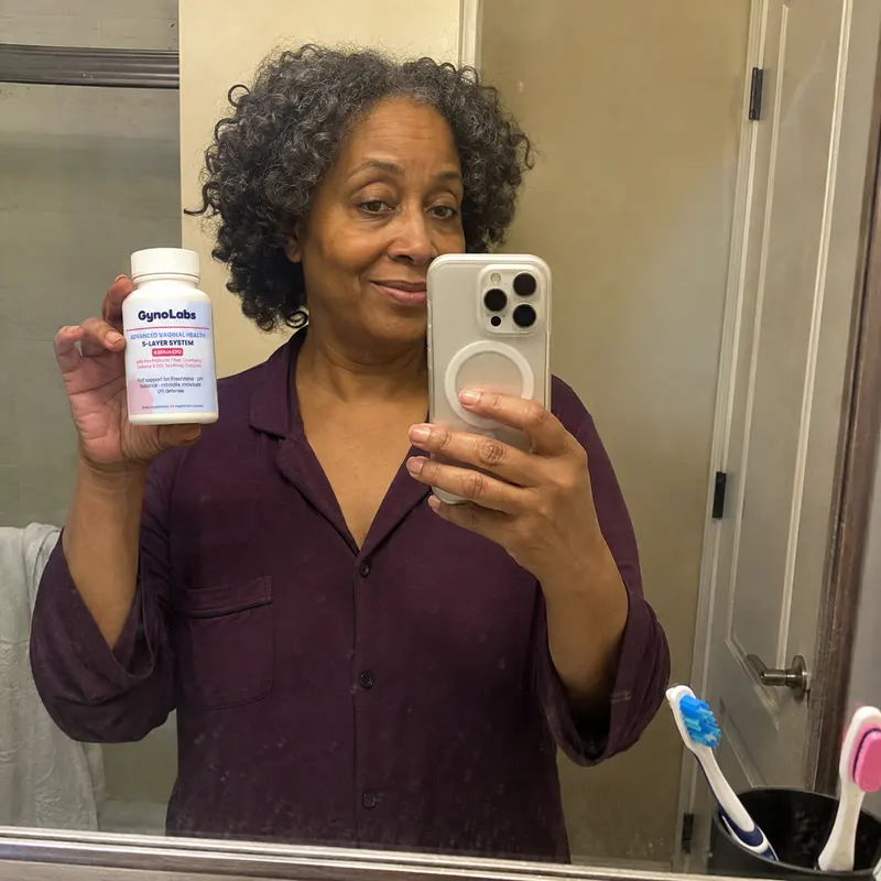 A woman with curly gray hair takes a mirror selfie while holding a bottle of GynoLabs supplements.