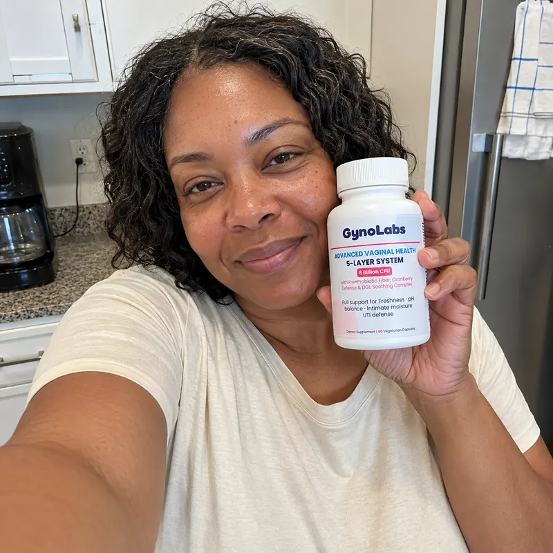 A woman with curly hair smiles while holding a bottle of GynoLabs supplements in her kitchen.