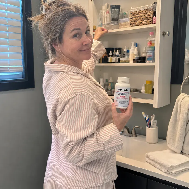 A woman in striped pajamas holds a bottle of supplements while reaching into her bathroom medicine cabinet.