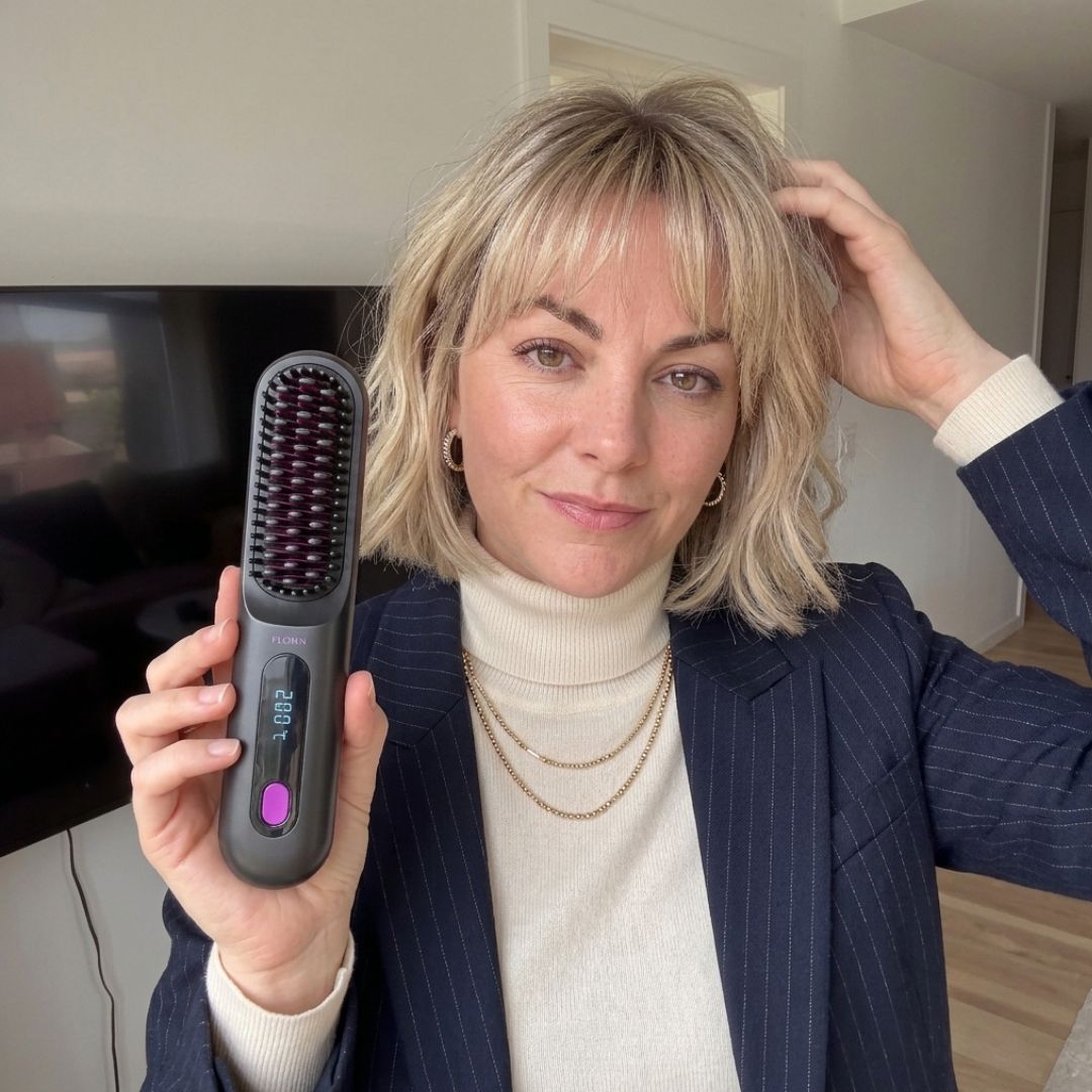 A woman with blonde hair holds a heated hairbrush in one hand and touches her hair with the other.