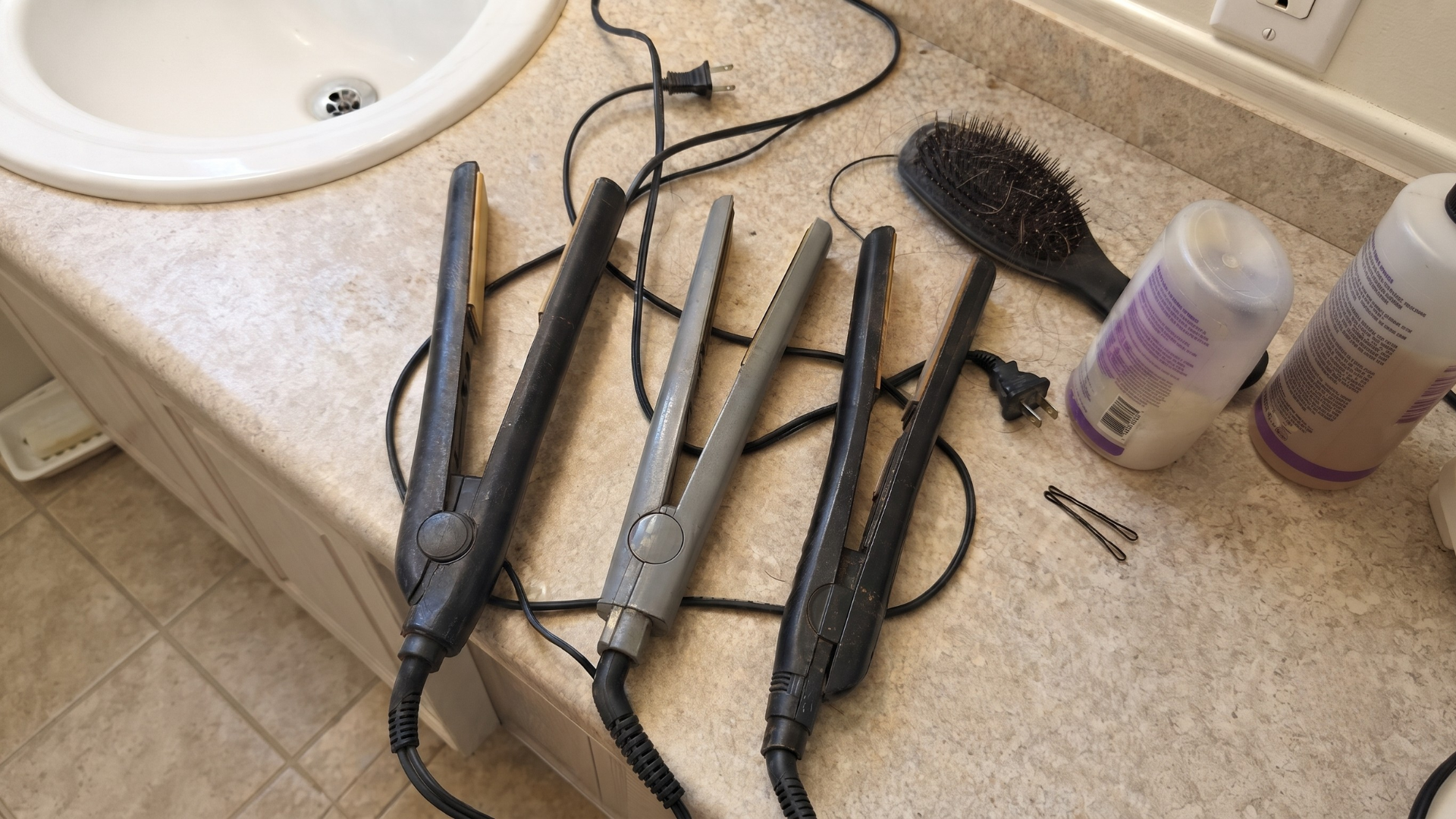 Three hair straighteners, a brush, and some hair products on a bathroom counter next to a sink.