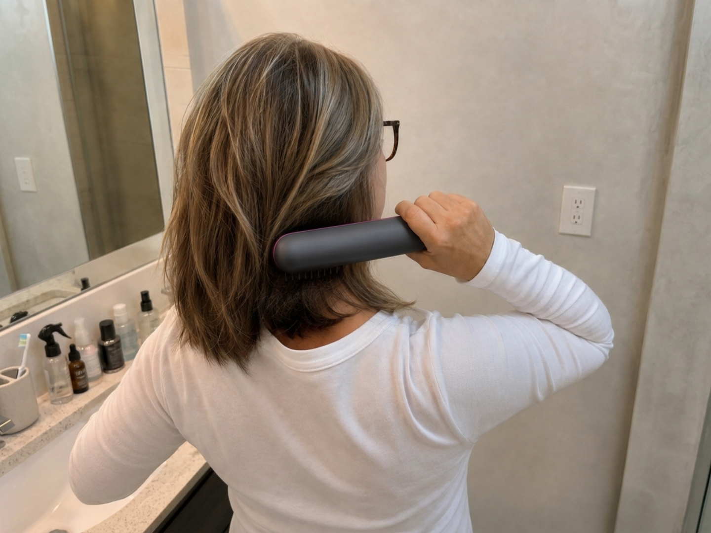 A person viewed from behind in a white shirt using a gray heated brush on their highlighted hair.