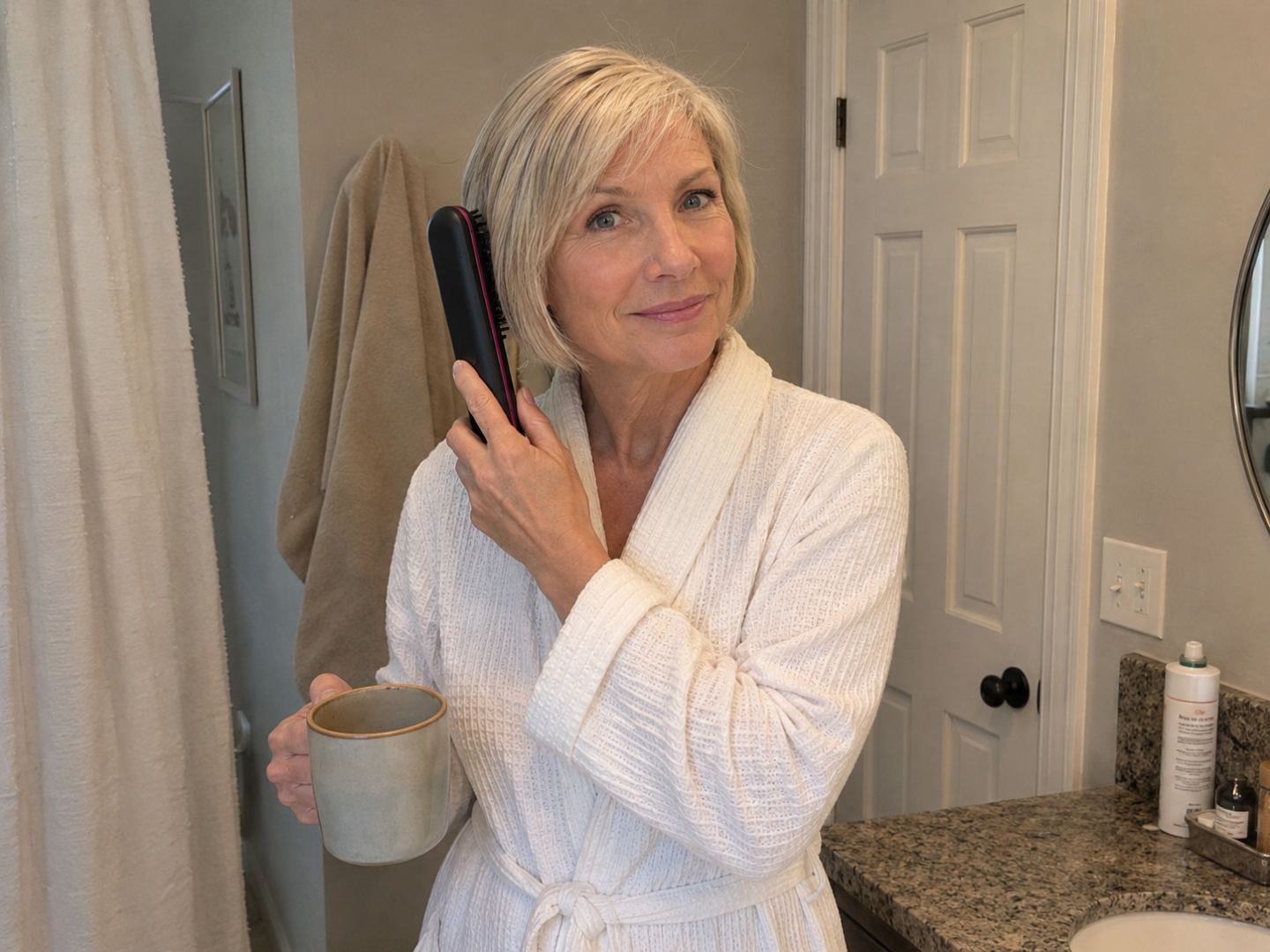 A woman in a white bathrobe stands in a bathroom, holding a mug and brushing her hair.