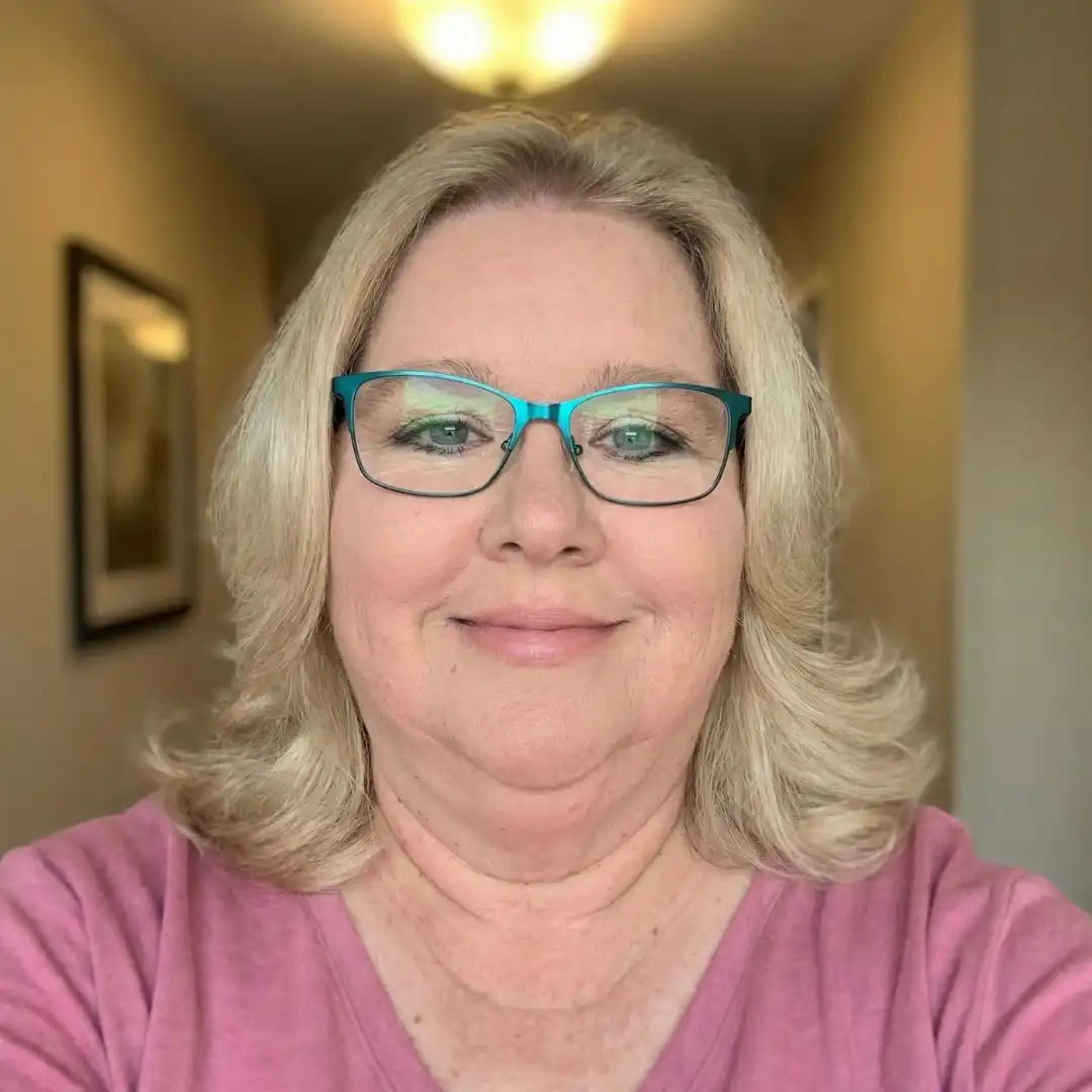 A smiling woman with blonde hair and teal glasses, wearing a pink shirt, looks at the camera.