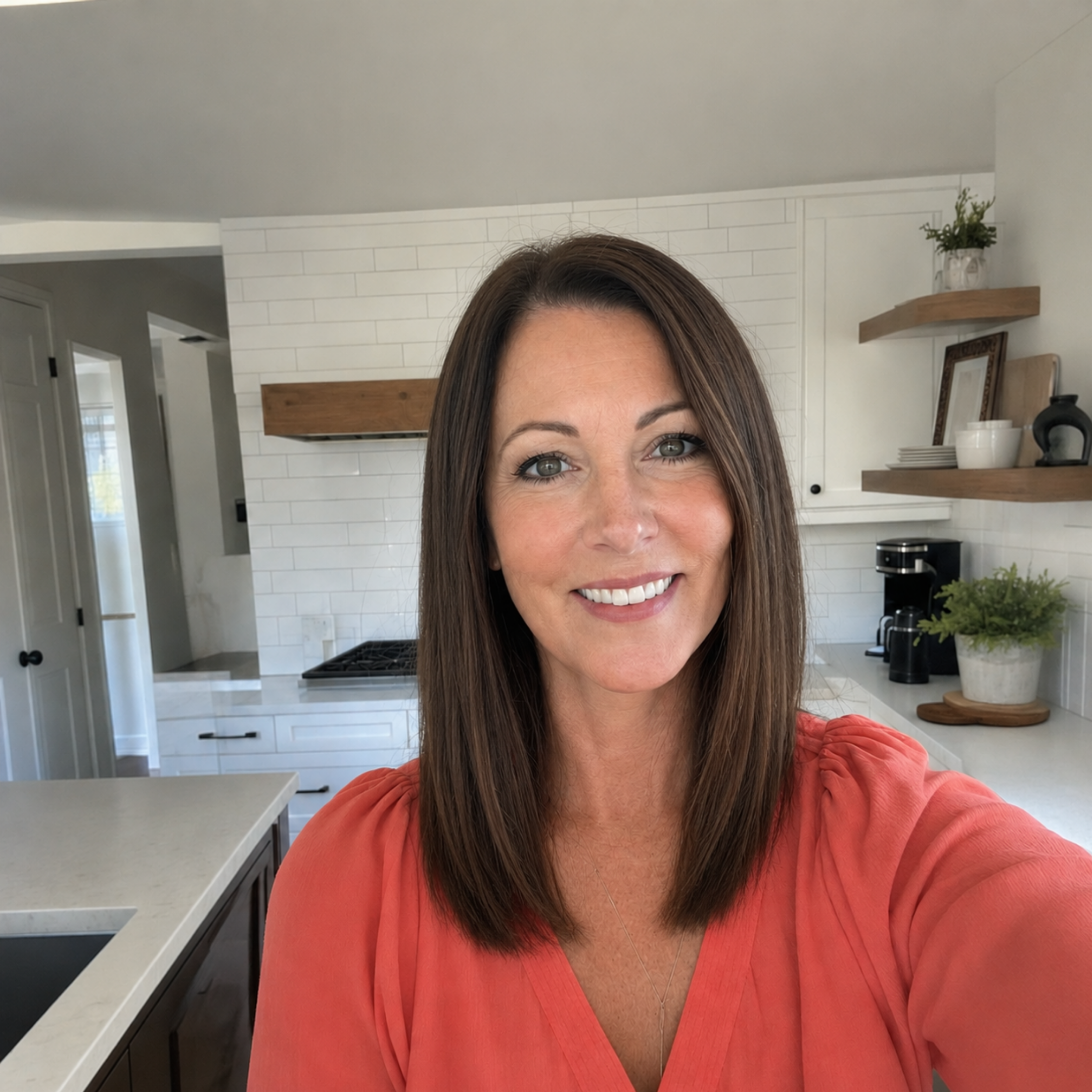 A smiling woman with brown hair and a coral shirt takes a selfie in a modern kitchen.