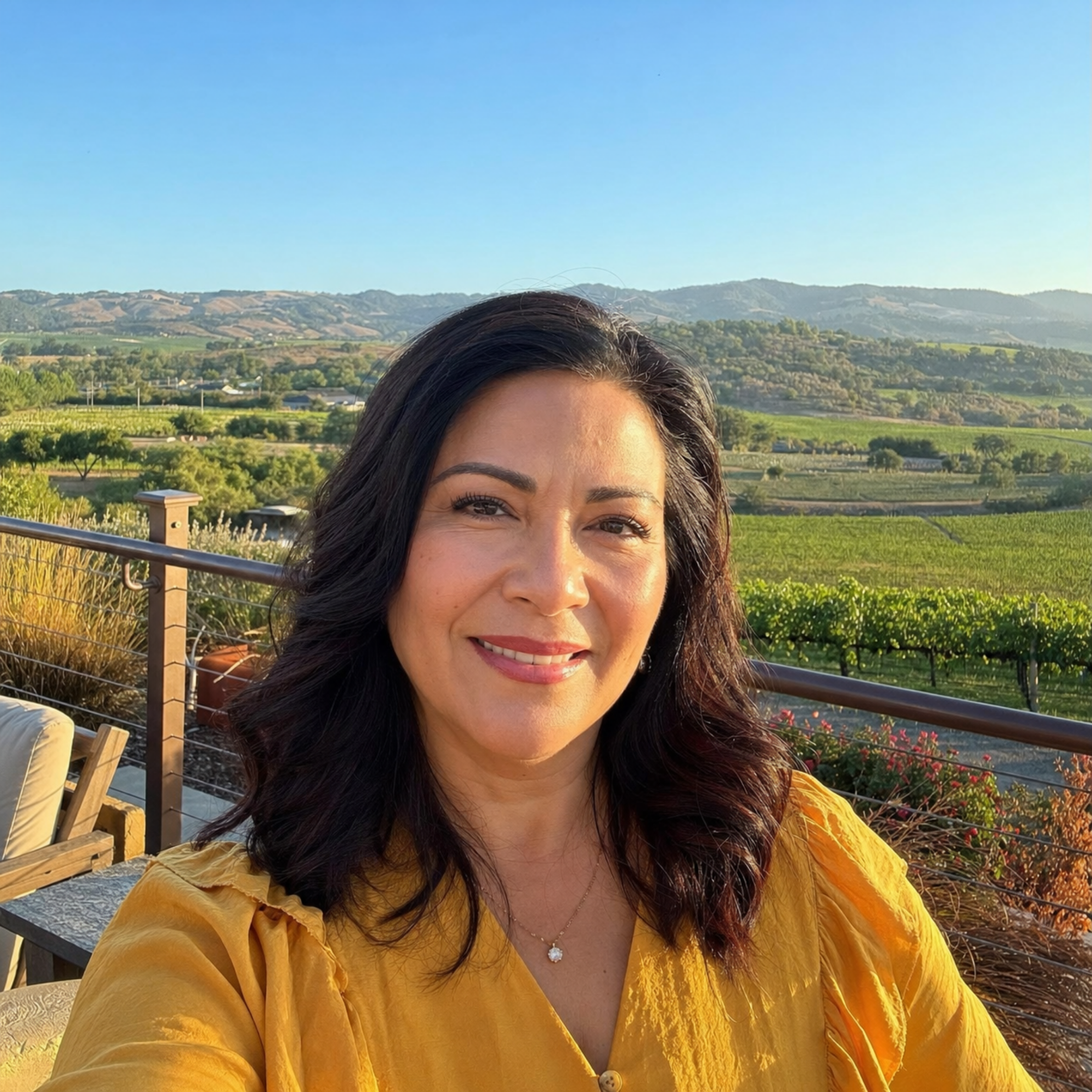 A woman in a yellow top smiles in front of a scenic vineyard and rolling hills.