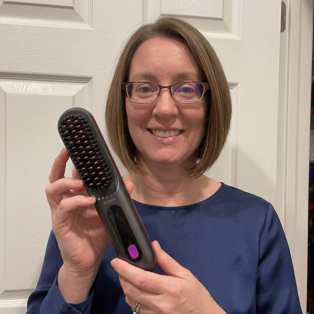 A smiling woman with glasses and short brown hair holds up a heated hair straightening brush.