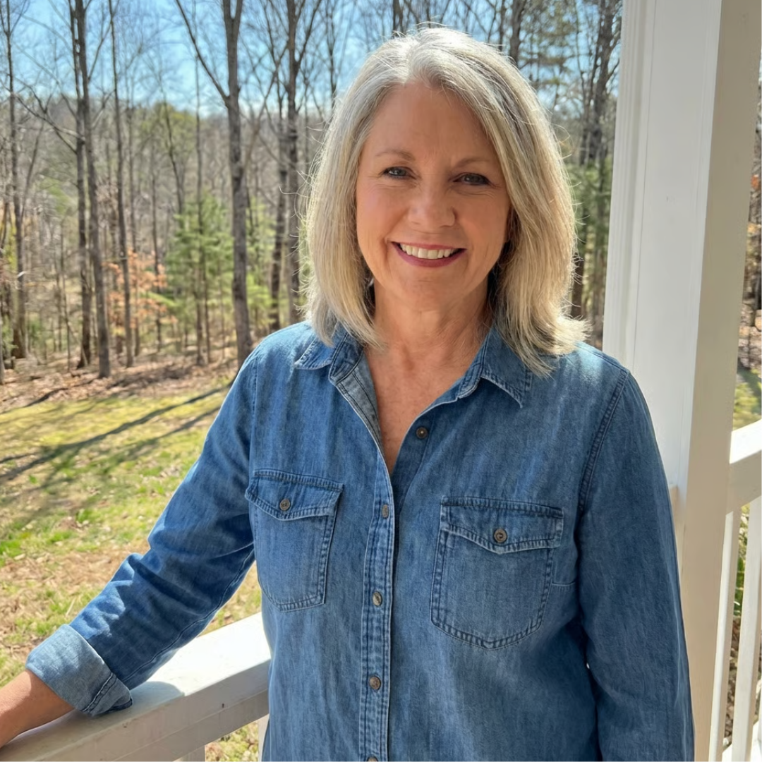 A smiling woman with shoulder-length gray hair and a denim shirt stands on a porch.