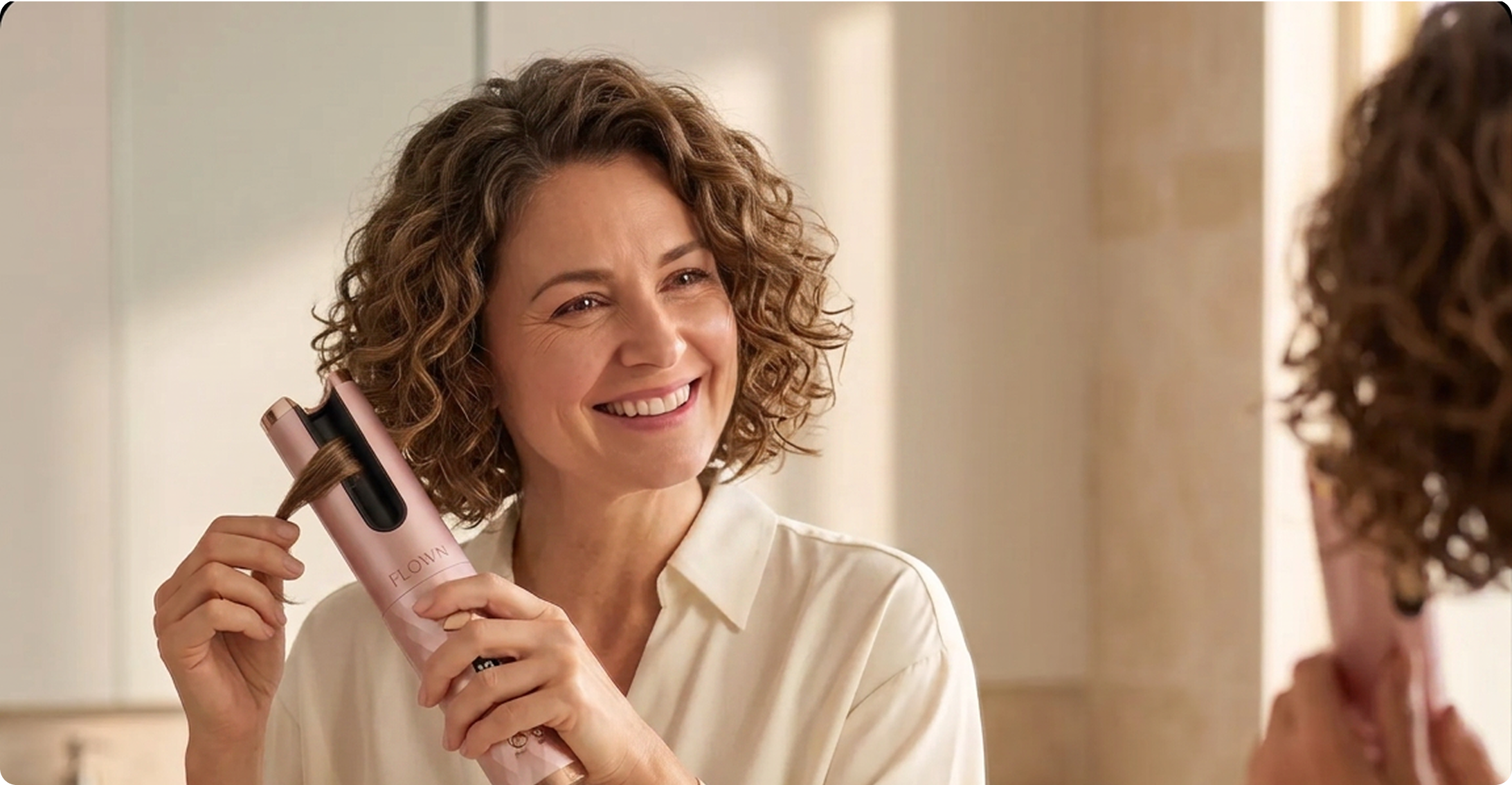 A smiling woman with curly hair uses a pink automatic hair curler in front of a bathroom mirror.