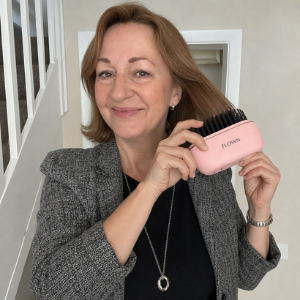 A smiling woman with reddish-brown hair holds a pink hairbrush to her hair.