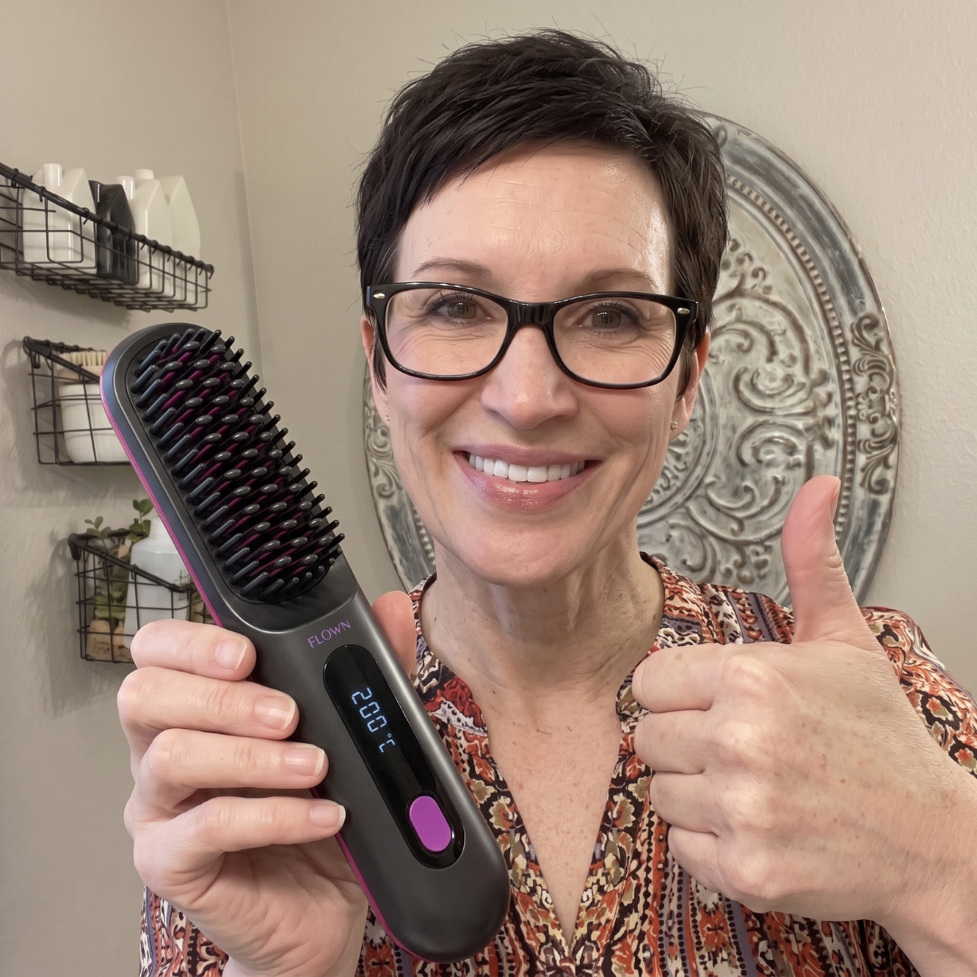 A smiling woman with glasses holds a heated hairbrush and gives a thumbs-up.