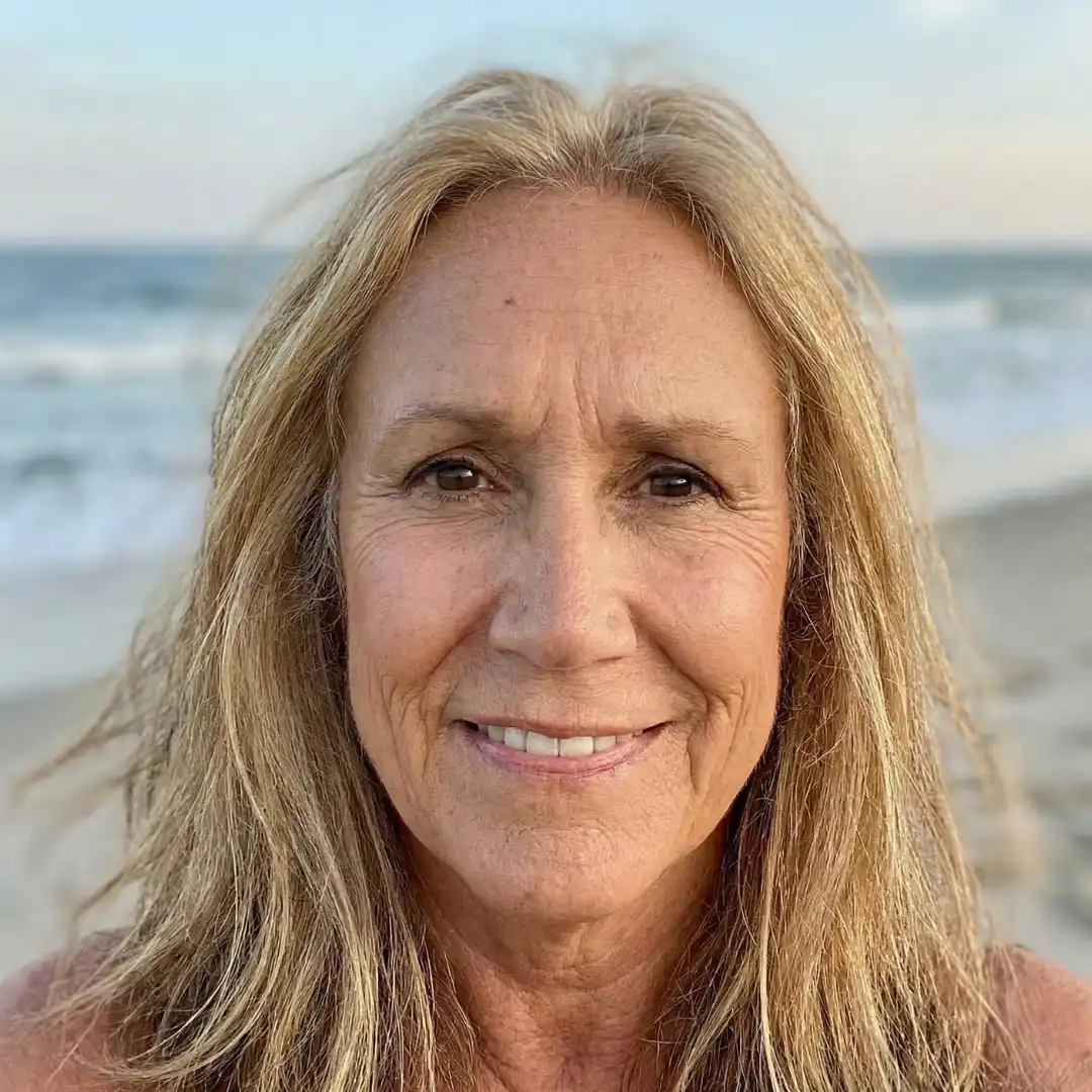 A close-up photo of a smiling woman with blonde hair, standing on a beach with the ocean behind her.