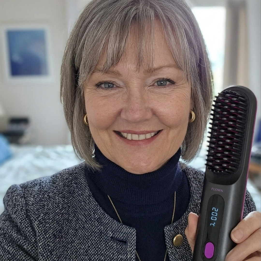 A smiling woman with short grey hair holds up a black heated styling brush.