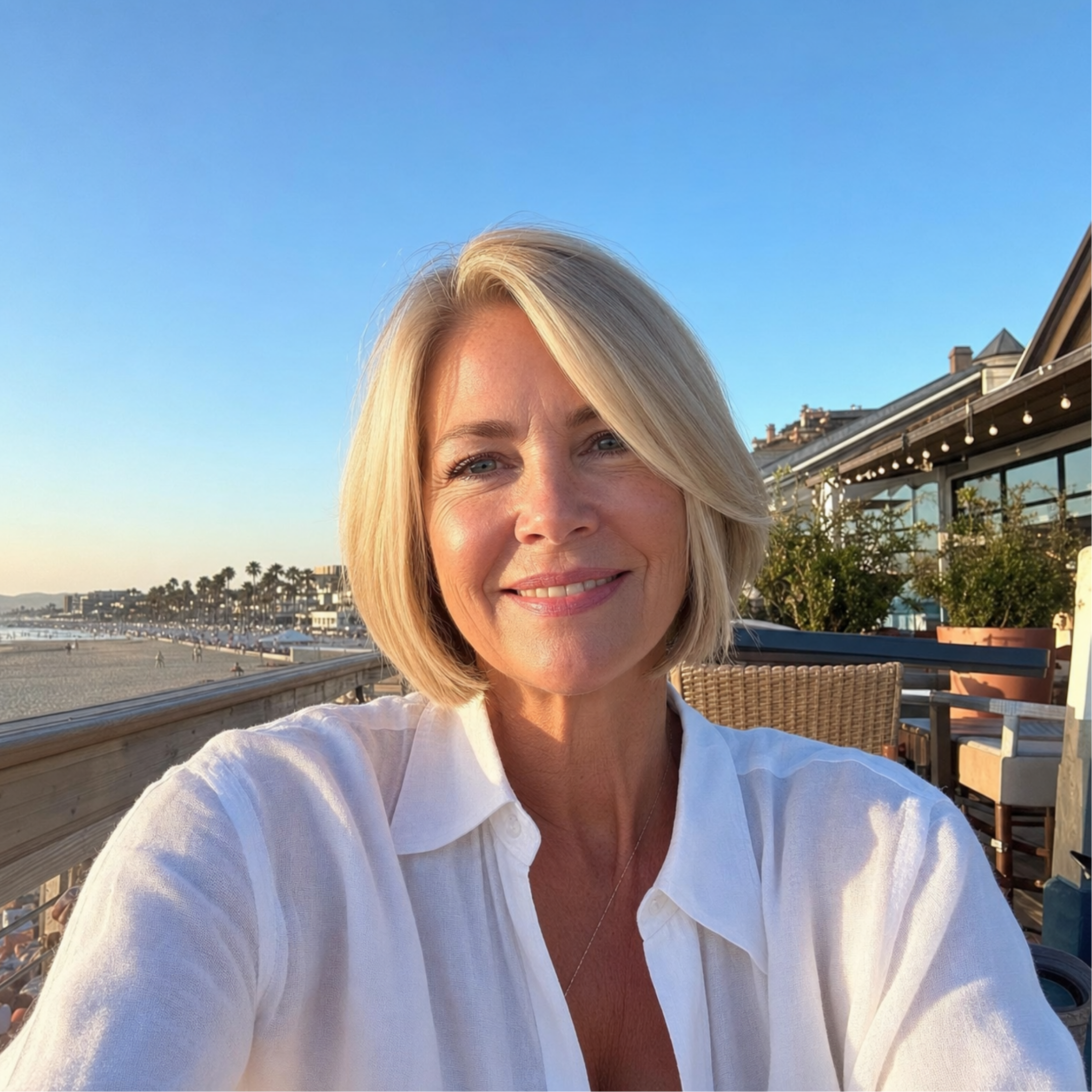 A smiling woman with short blonde hair takes a selfie on a patio overlooking a beach.