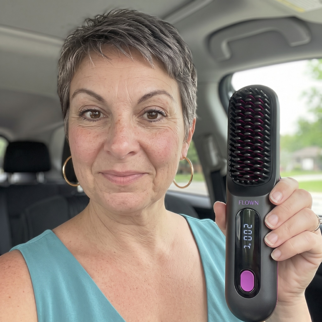 A woman with short hair sits in a car, holding up a heated hair straightening brush.