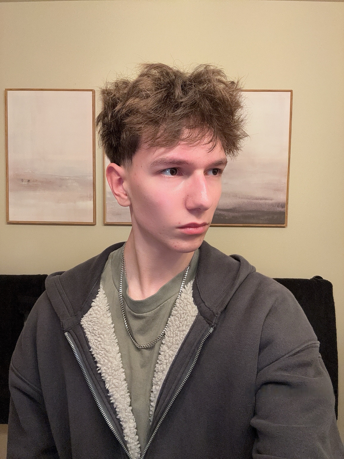 Person with short hair wearing a jacket, standing indoors with framed art on the wall.