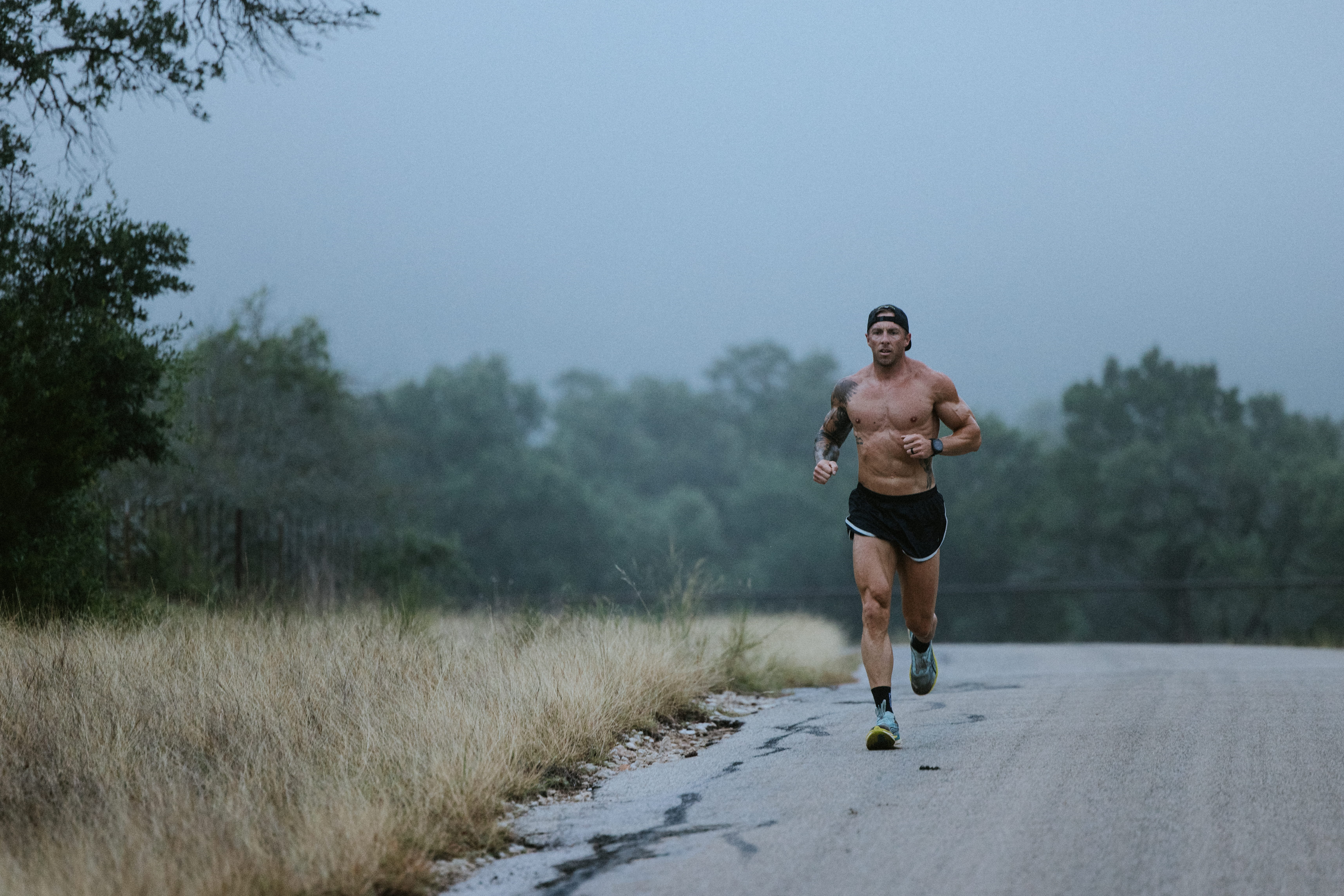 A shirtless, muscular man with tattoos runs on a rural road on a foggy day.