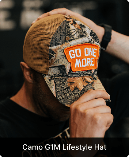 A person adjusts a camouflage trucker hat with an orange patch that reads 'GO ONE MORE'.