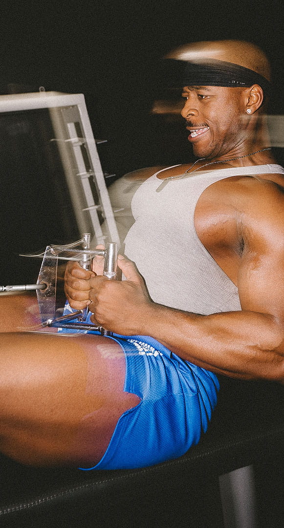 A muscular man in a tank top smiles while using a leg exercise machine at the gym.