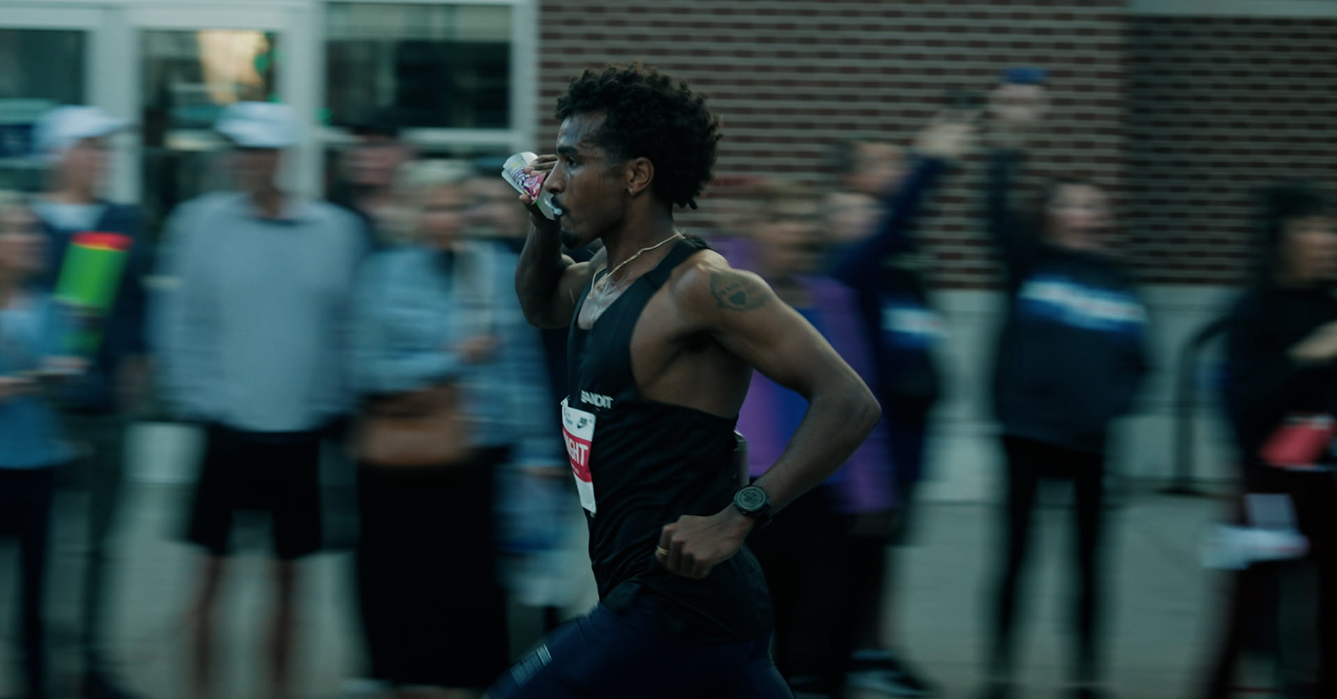 A male runner in a black tank top drinks from a bottle while running during a race.