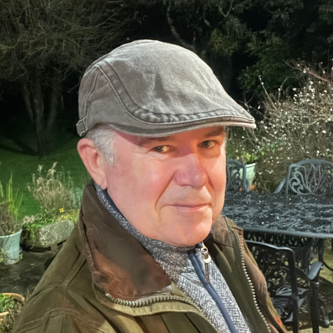 A man in a grey flat cap and brown jacket sits outdoors at night, looking towards the camera.