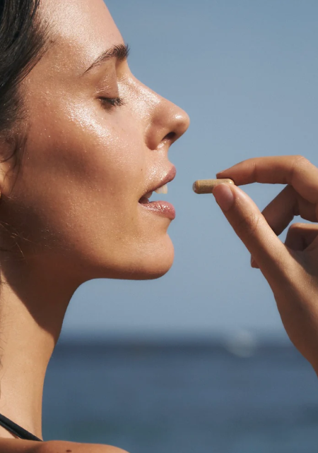 A close-up profile of a woman with her eyes closed, holding a tan capsule to her open mouth.