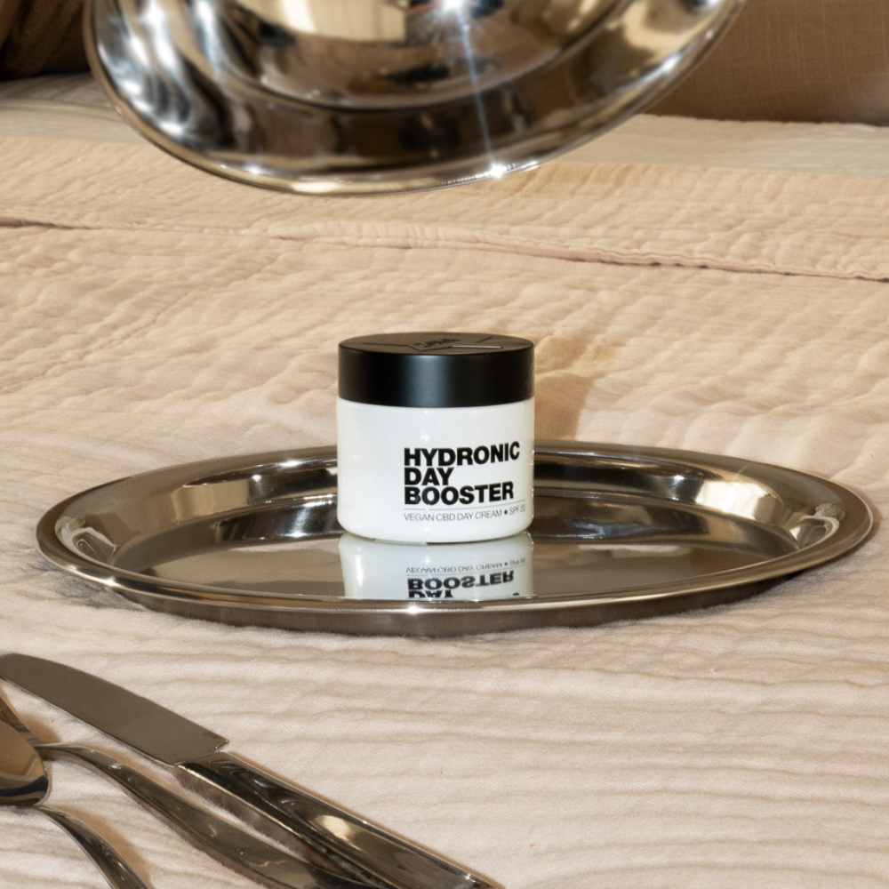 A jar of Hydronic Day Booster cream rests on a silver tray set on a rumpled pink cloth.