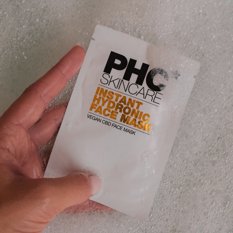 A hand holds a white sachet of PHC Skincare face mask against a background of white foam.