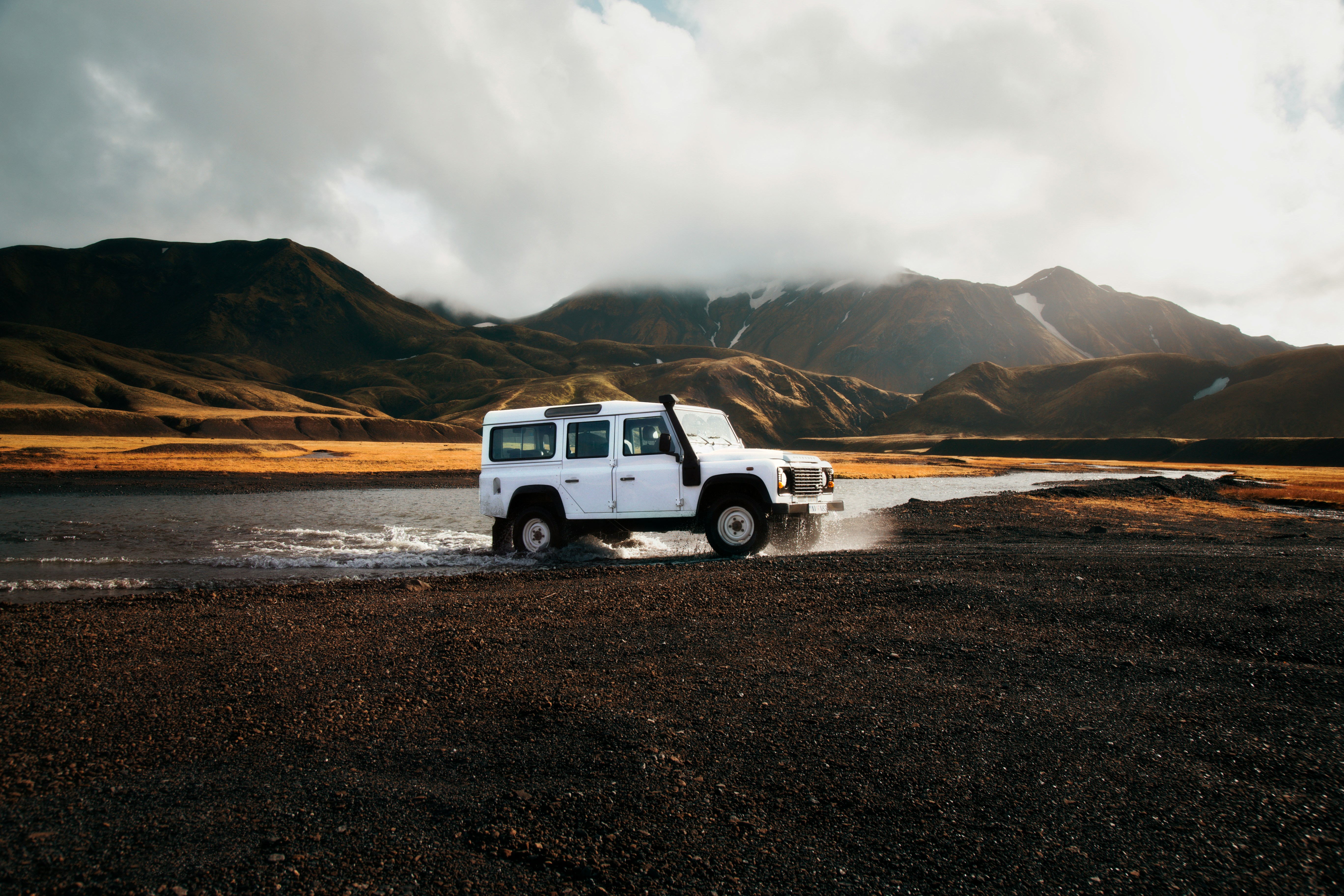 A white off-road vehicle drives through a shallow river with mountains in the background under a cloudy sky.