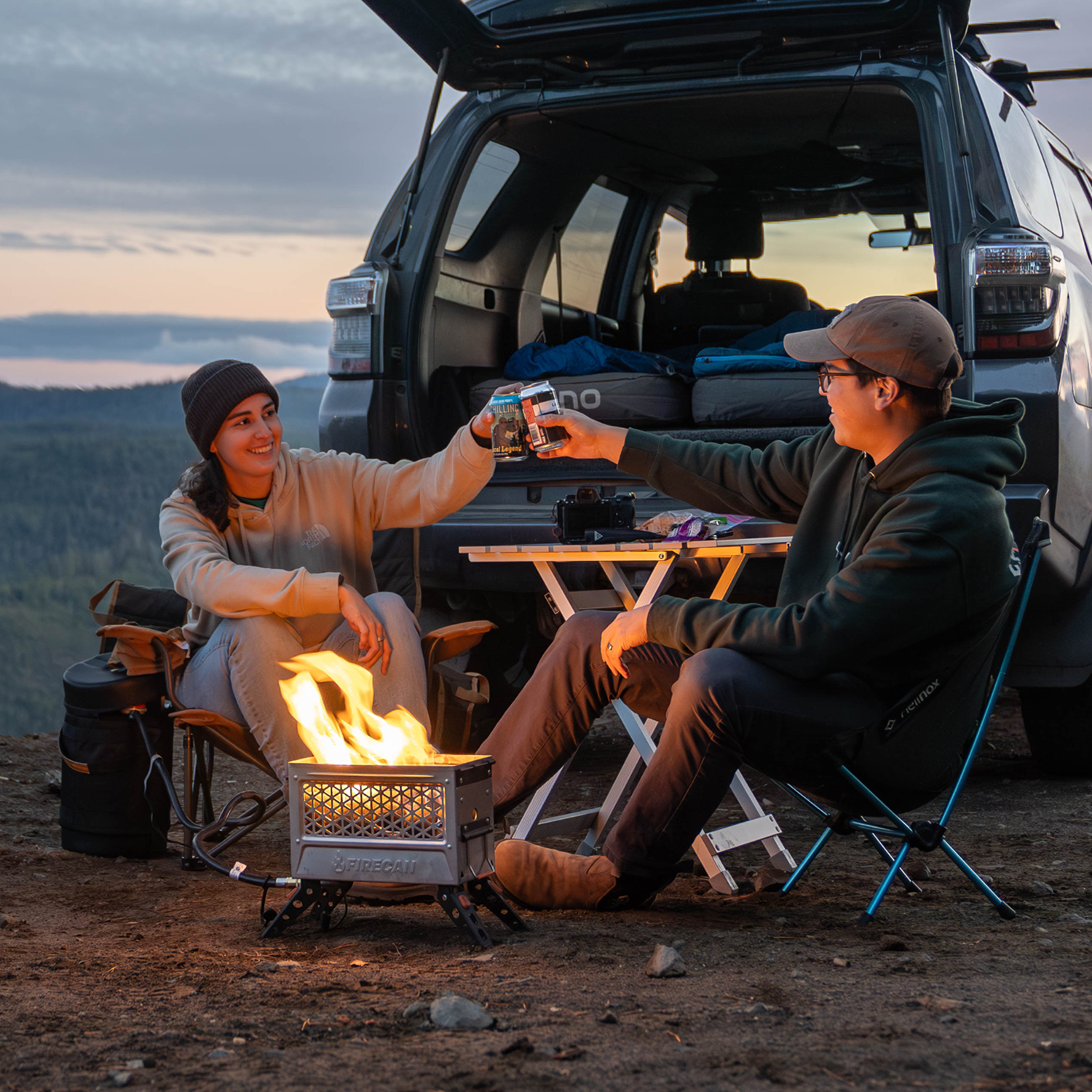 Two people camping, sitting by a fire, toasting drinks near an open car trunk.