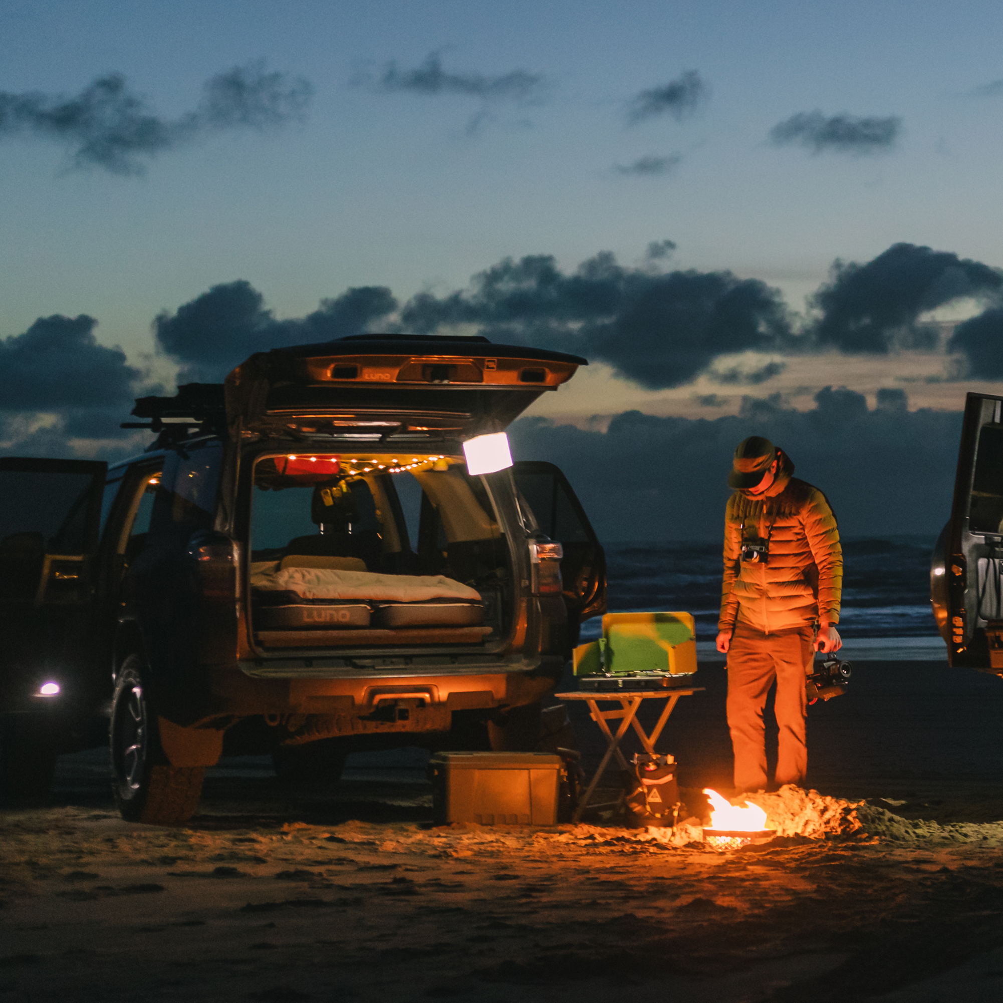 A person stands by a campfire on a beach at dusk next to an SUV.
