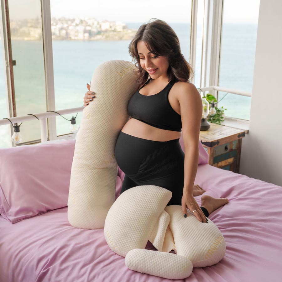 Pregnant woman in black outfit sitting on bed with a large body pillow.