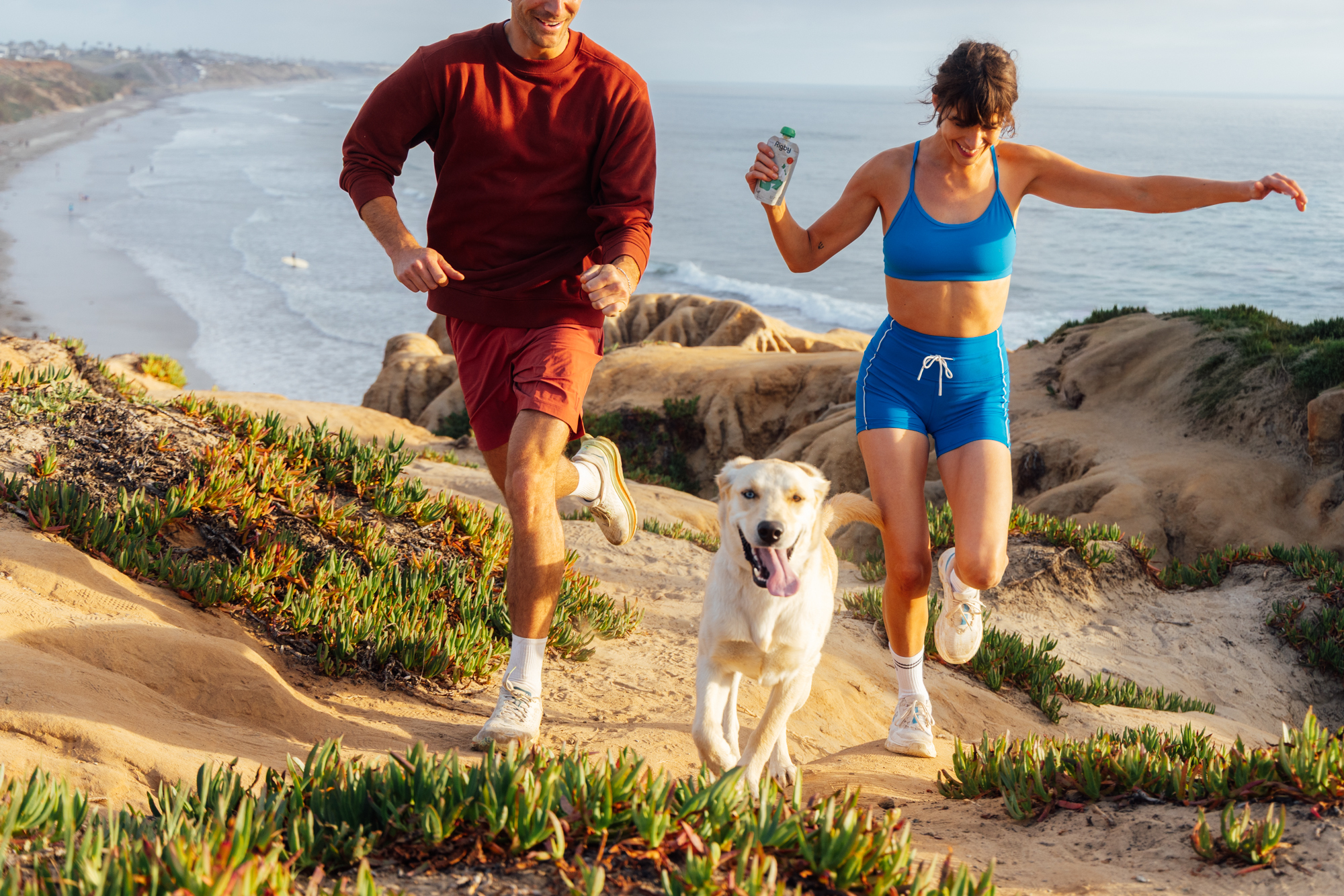 A man, a woman, and a dog run along a dirt path on a cliff overlooking the ocean.