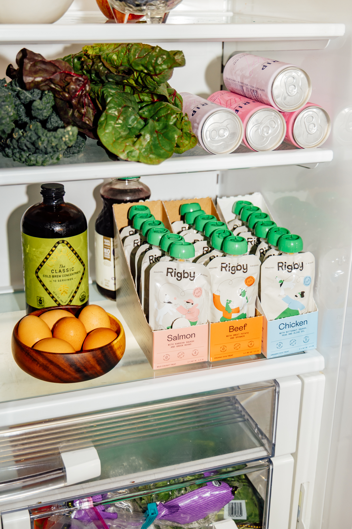 An open refrigerator stocked with fresh produce, eggs, canned drinks, and Rigby brand dog food pouches.