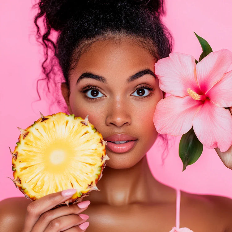 Person holding a pineapple slice and pink hibiscus flower against a pink background.