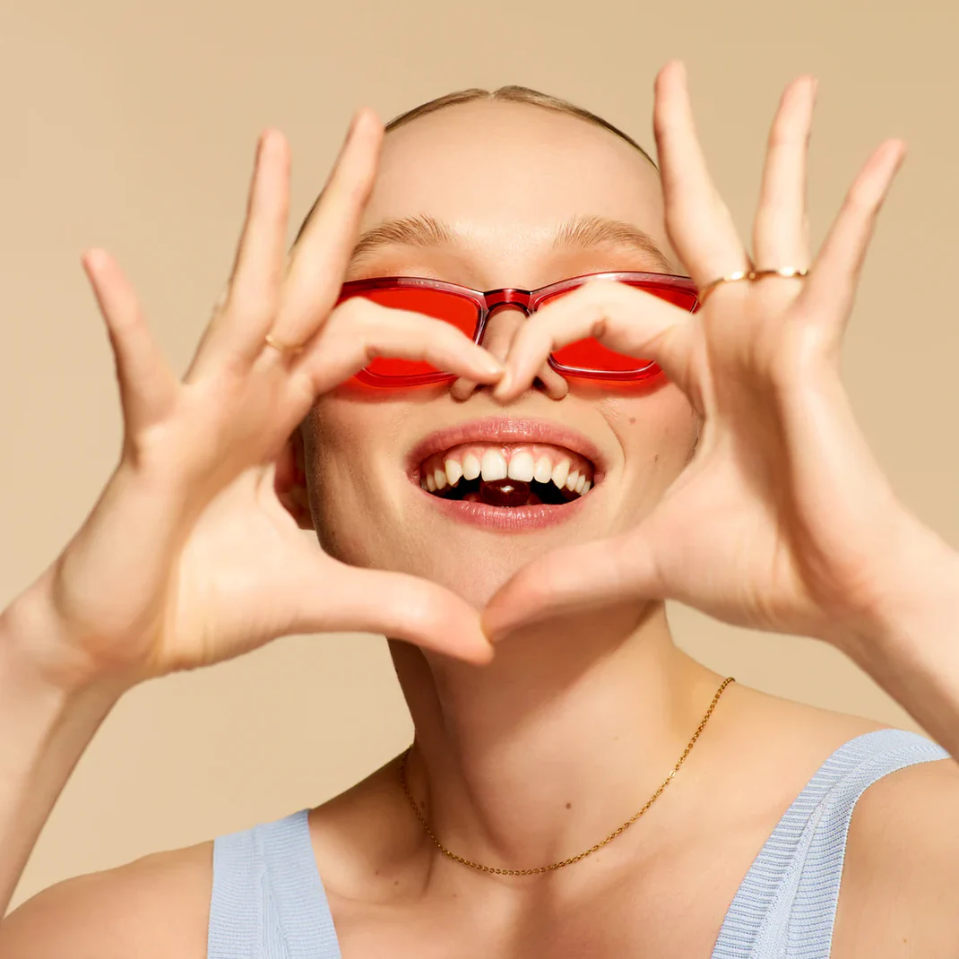 Person smiling with red sunglasses forming a heart with hands.