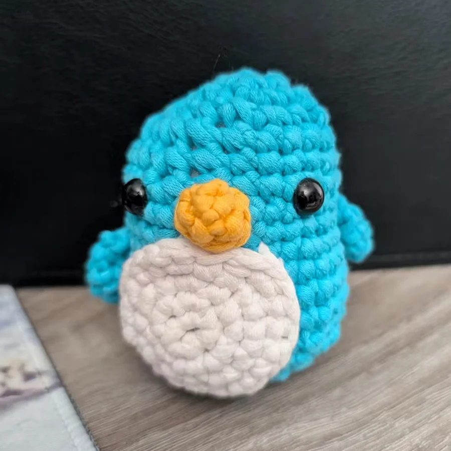 Crocheted blue penguin with orange beak and white belly on a wooden surface.
