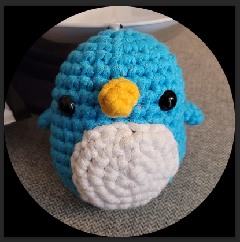 Crocheted blue penguin plush with a yellow beak and black eyes on a gray surface.