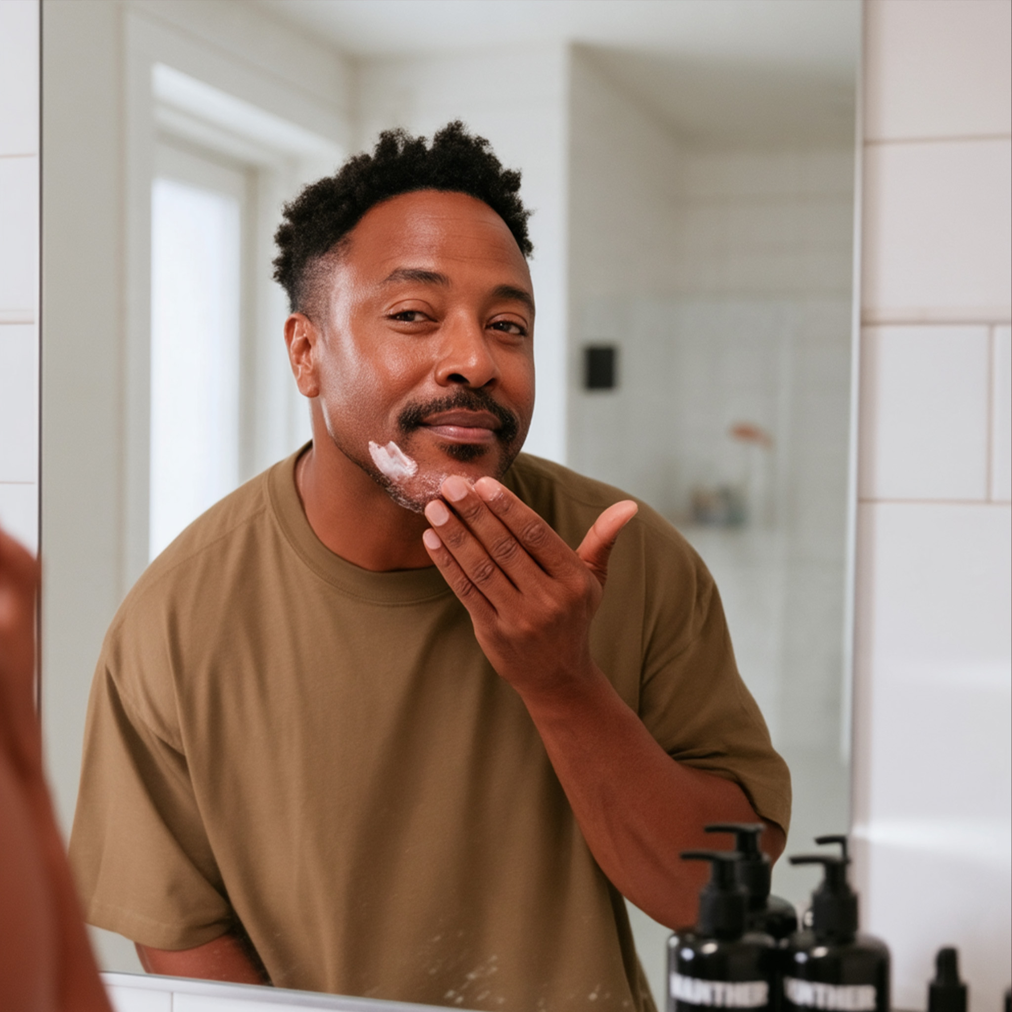 A man in a brown t-shirt applies cream to his chin while looking in a bathroom mirror.
