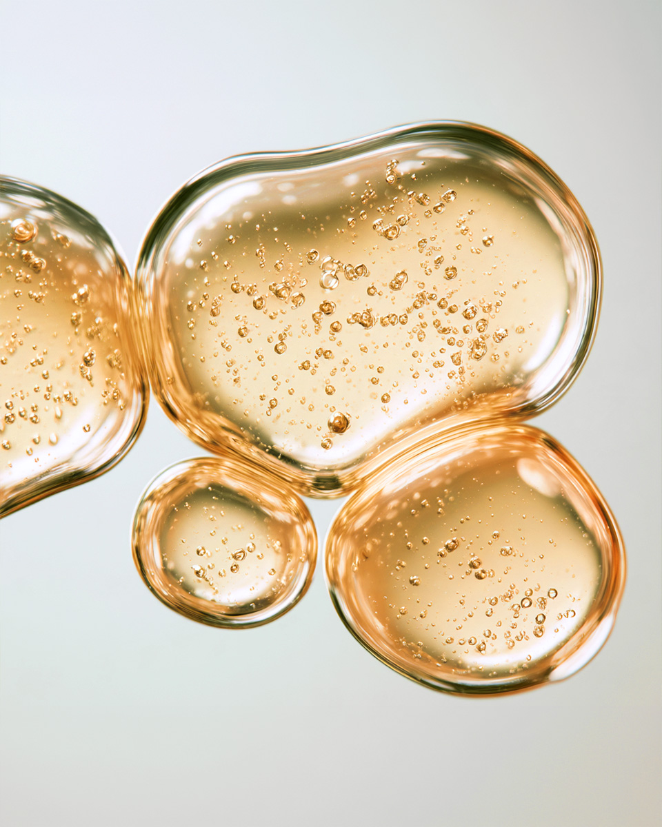 A close-up of several drops of a golden, bubbly liquid on a light gray background.