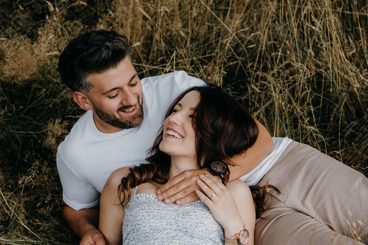 A smiling couple lies together in a field of tall grass, looking at each other.
