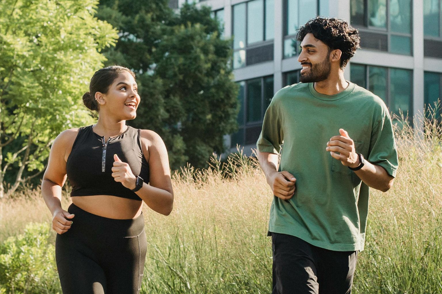 A man and a woman in athletic clothes smile at each other while jogging through a grassy area.