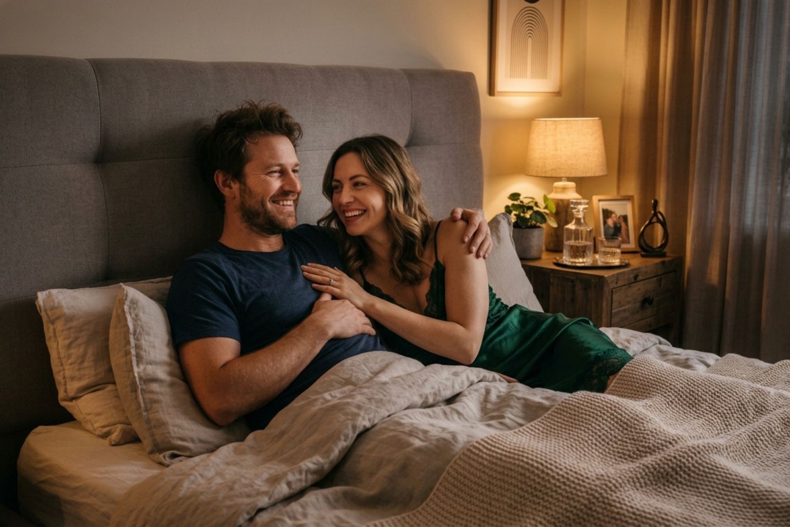 A smiling man and woman cuddling together in a cozy bed at night.