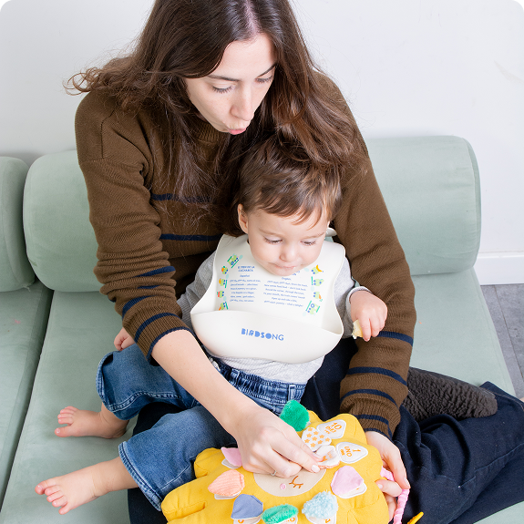 A woman and a toddler sit on a couch, looking down and playing with a yellow toy.