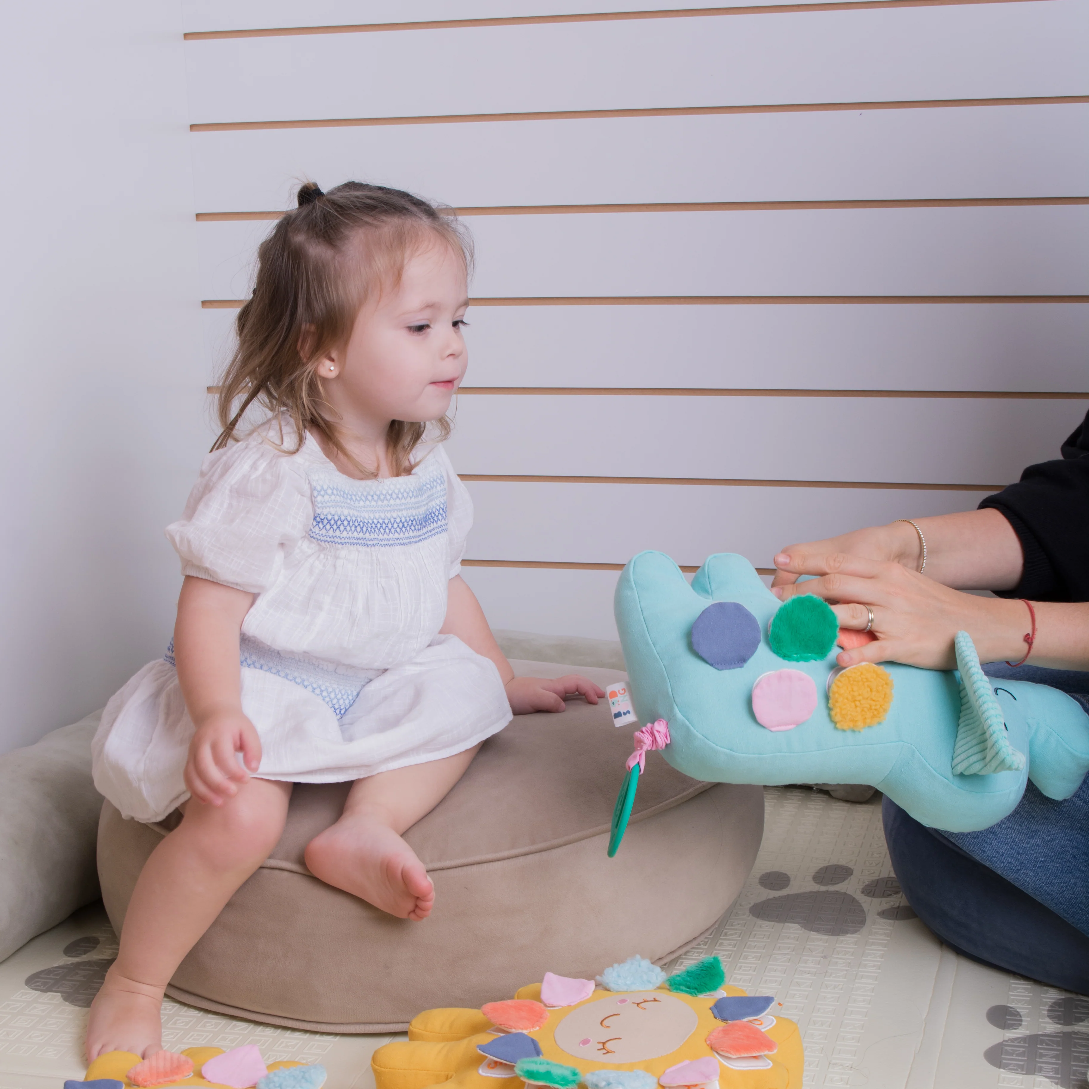 A little girl in a white dress sits on a cushion playing with an adult and a stuffed toy.