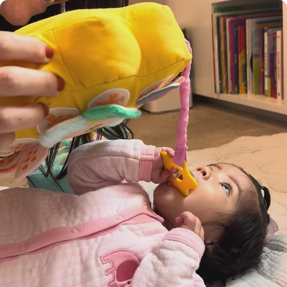 A baby lies on the floor, chewing on a teether while looking up at a plush toy.