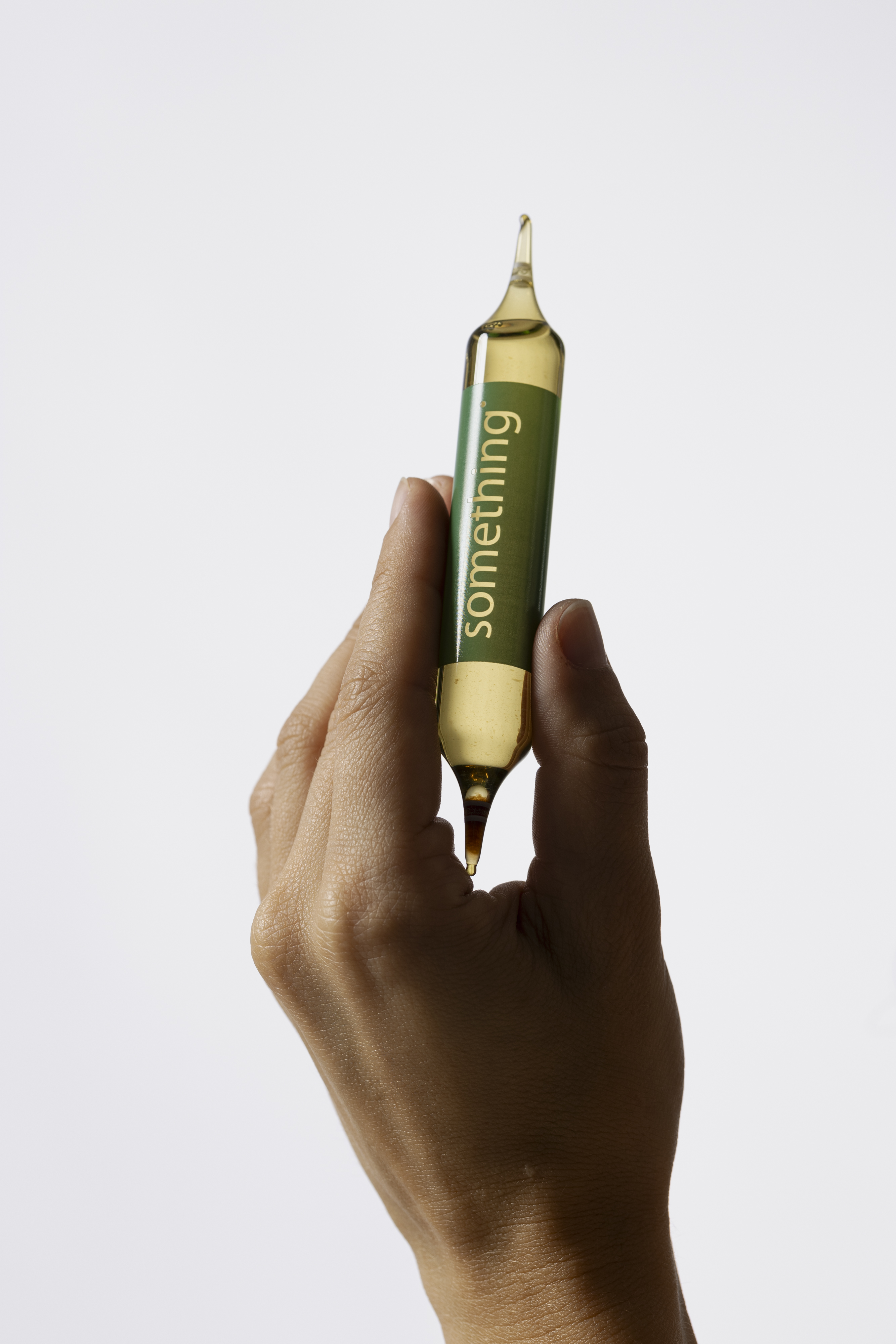 A hand holds a glass ampoule with a yellow liquid and a green label reading 'something'.