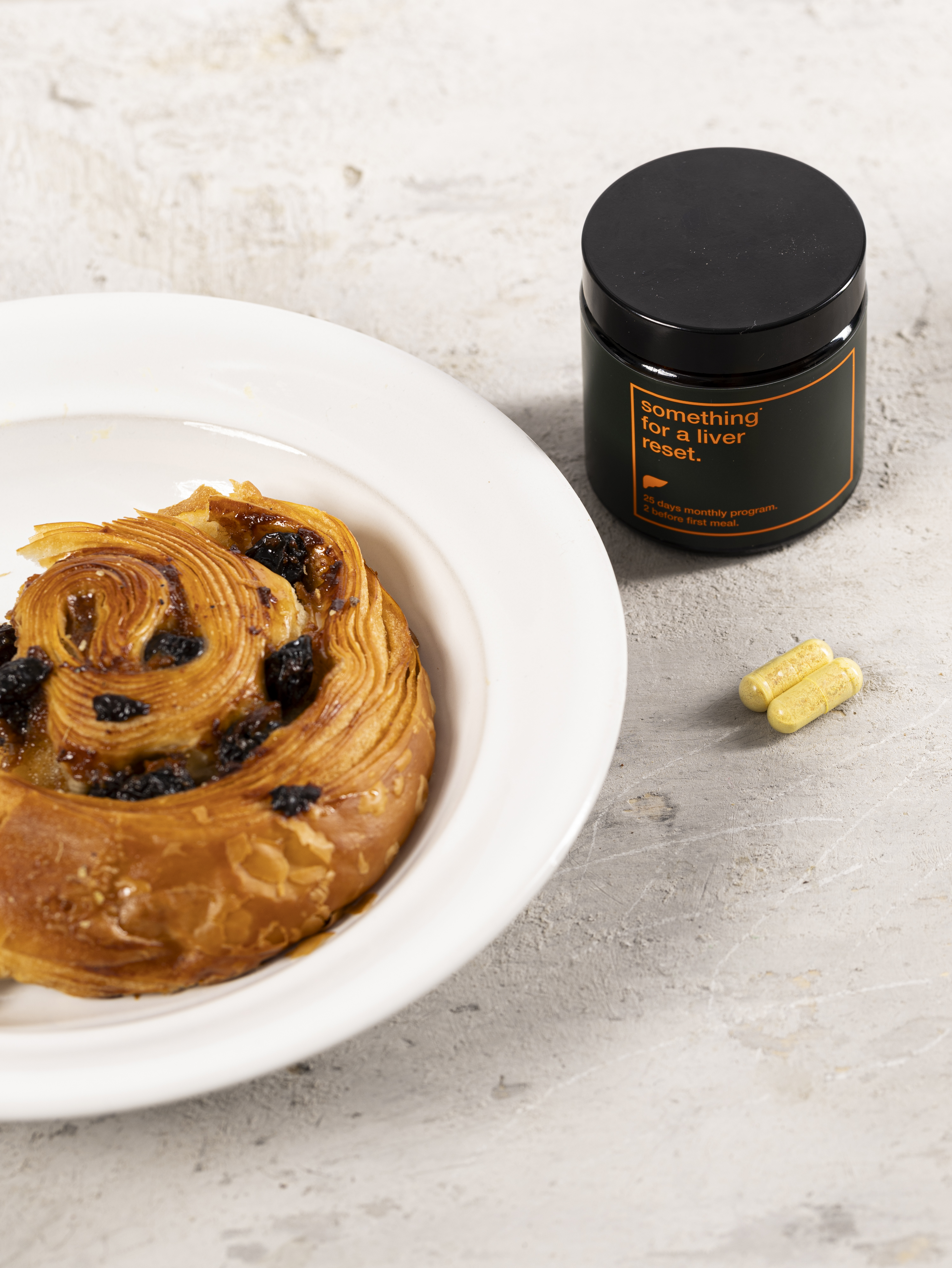 Pastry on a plate, jar labeled 'liver reset,' and two capsules on a table.