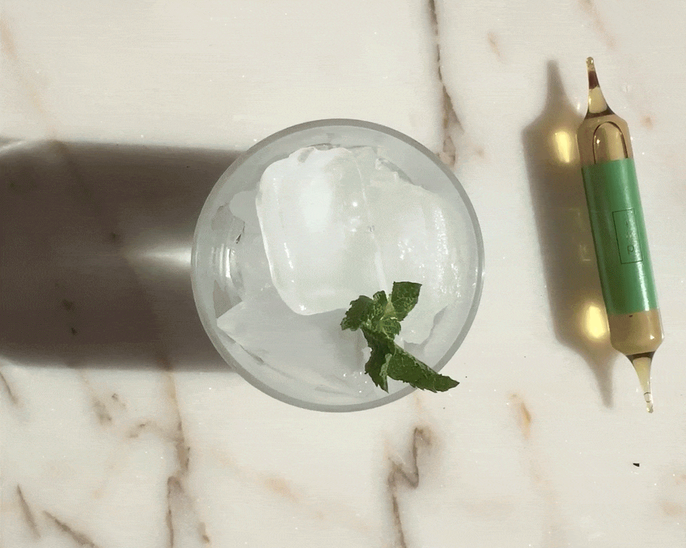 Glass with ice and mint next to a green ampoule on a marble surface.