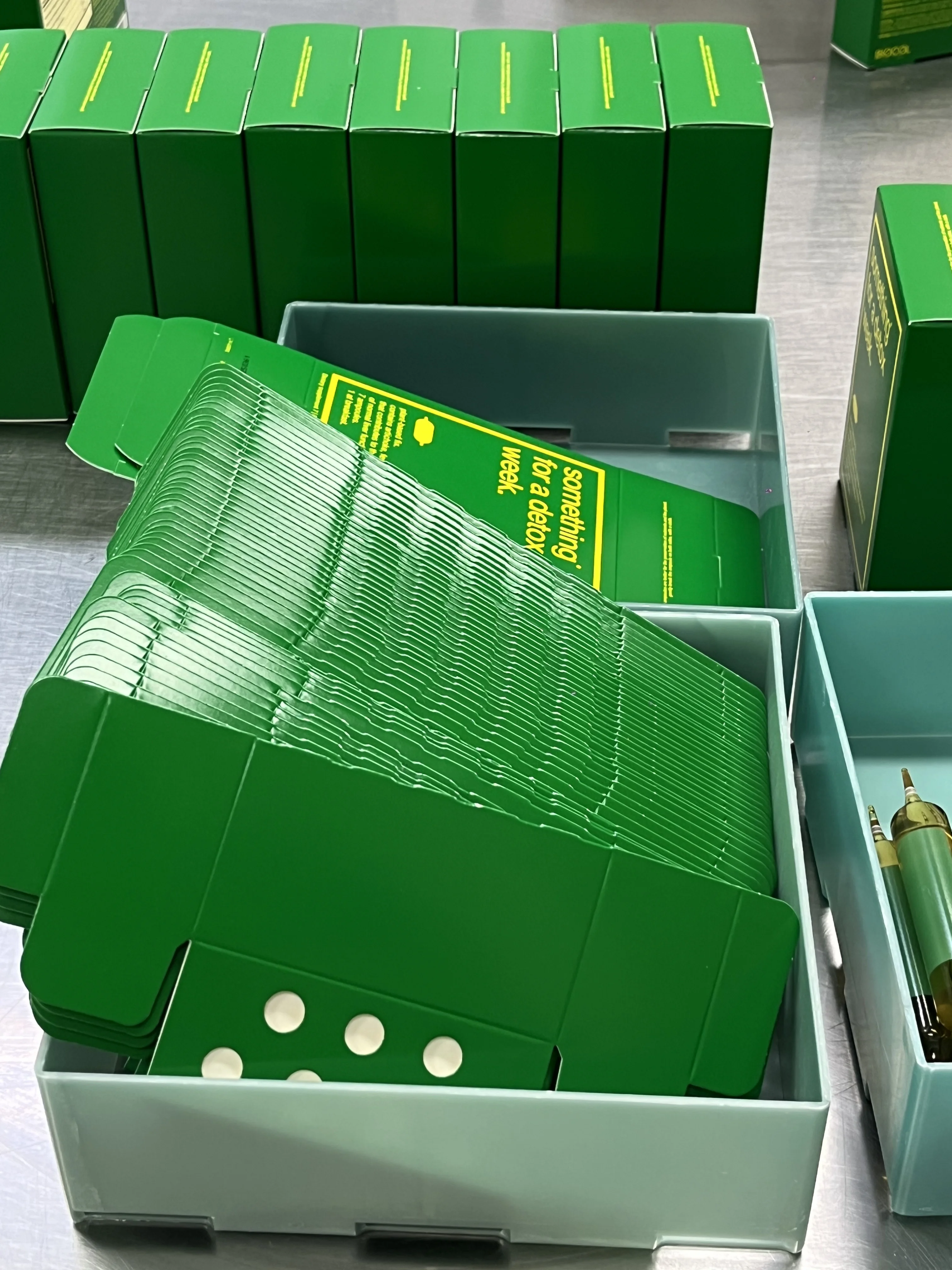 Stacks of unfolded green boxes in trays, with assembled boxes and glass ampoules nearby on a table.